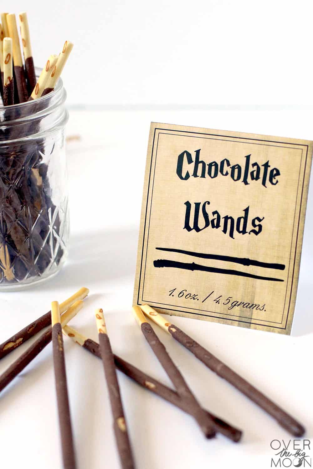 Some chocolate wafer sticks in a clear jar and laying on a white table. Next to them is a candy label that says Chocolate Wands.